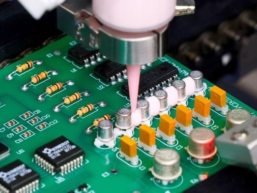 Nahaufnahme der automatisierten Dosierung eines dickflüssigen, hellrosafarbigen Beschichtungsmaterials um silberne Komponenten auf einer grünen Leiterplatte